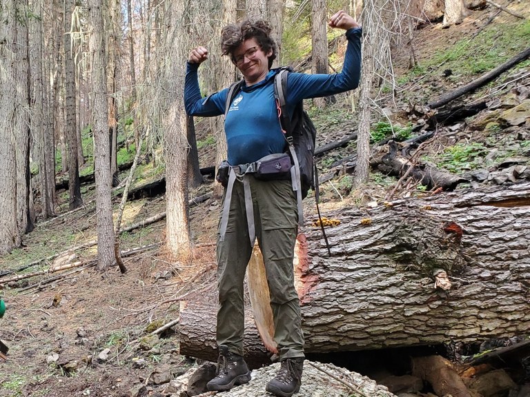 Rhoda has a wealth of trail work experience from across the country and can't wait to test her strength this season. Photo by Rhoda Boettcher A trail worker stands upon a felled log. They flex their biceps in triumph.