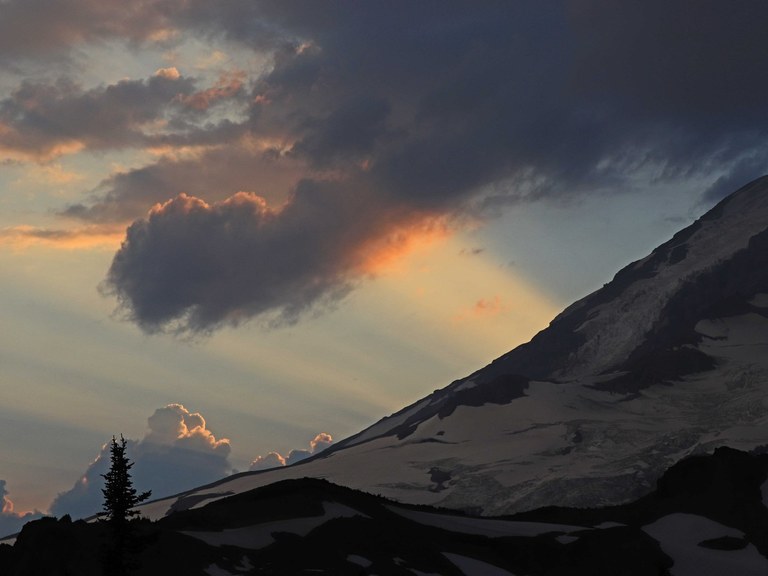 A dramatic mountain slope is in the foreground, with clouds and sunshine streaking through the sky in the background against a gray sky.