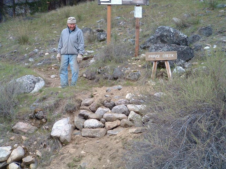 A volunteer in jeans and a fleece looks proudly at a rock wall on trail