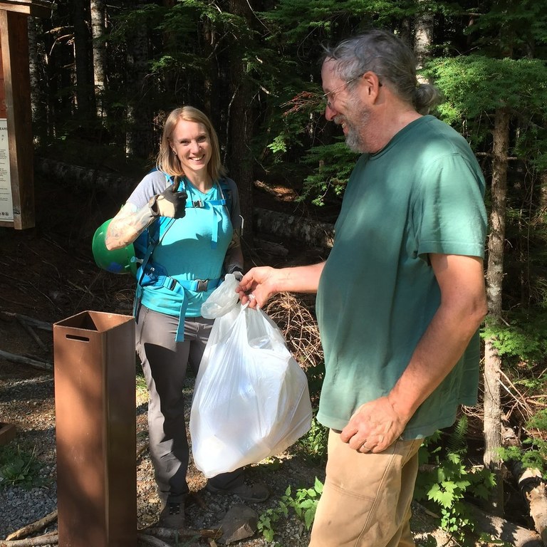 Two hikers are holding a trash bag at the trailhead. One smiles at the camera and offers a thumbs up while the other holds the bag. 