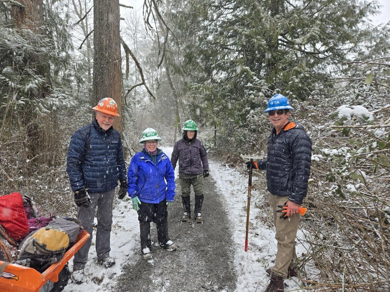 Vols smiling in the snow at Ridgefield national wildlife refuge Four people stand on a gravel trail with snow falling around them. They hold tools and smile at the camera.
