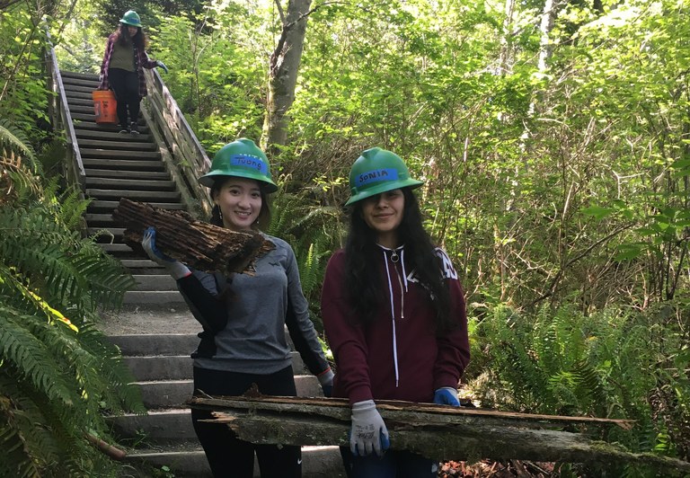 Two youth volunteers stand at the base of a staircase in Discovery Park. They are smiling and holding some wood they have removed from the trail. Photo by Kaci Darsow.
